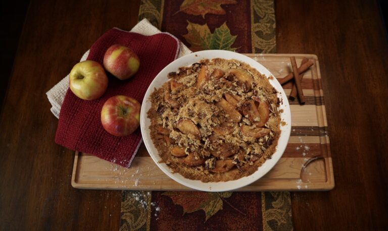 A classic apple pie with pecan crumbles sitting on a multi-colored cutting board with apples and cinnamon sticks and old-fashioned towels